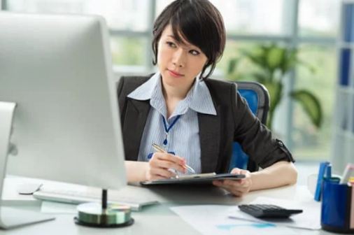 A worker sitting on a chair, looking at a computer on the desk, and taking notes.